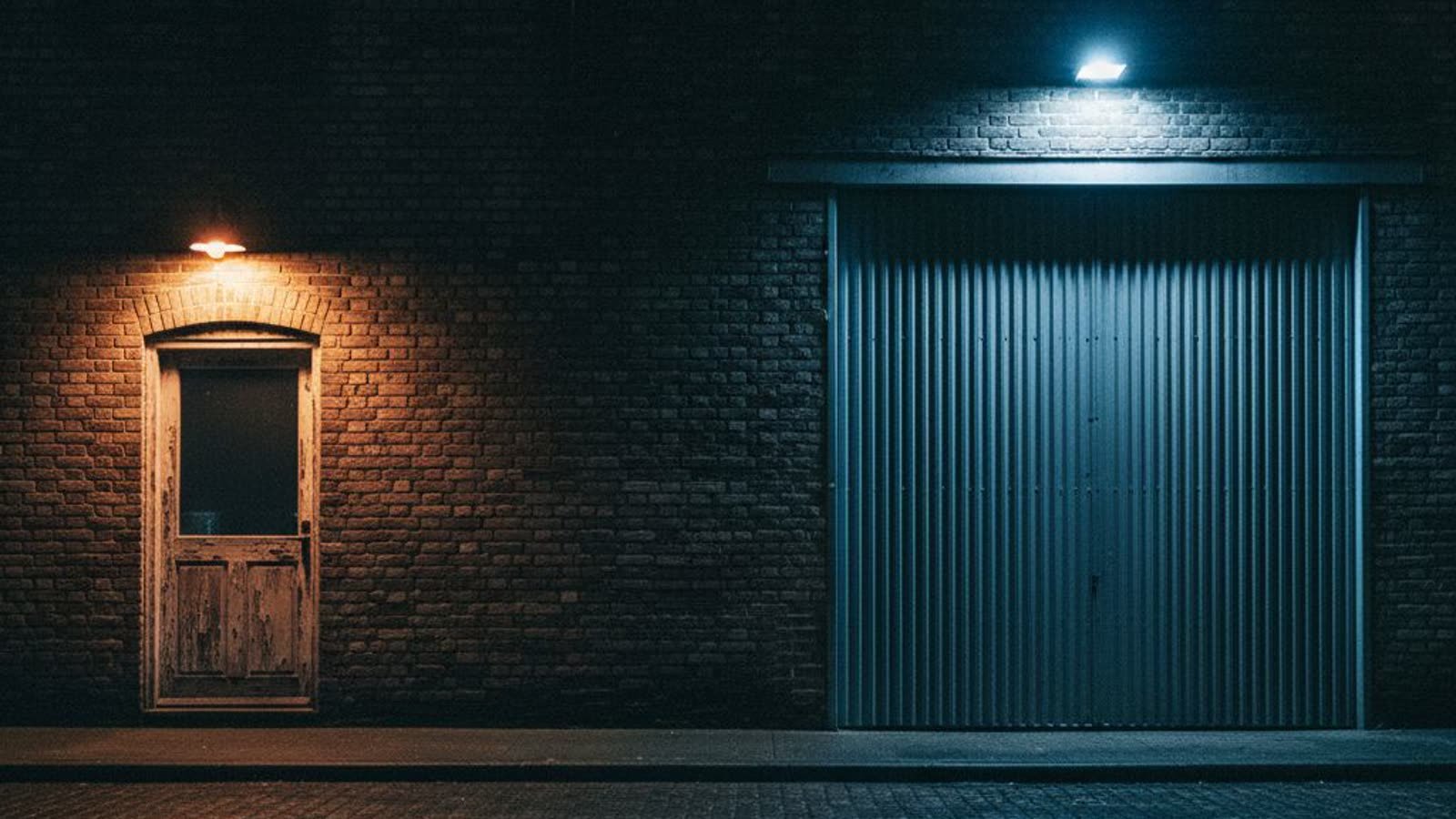 A small wooden shop door lit by a warm lamp and a large steel loading door under cold floodlight, side by side in one brick wall at night.