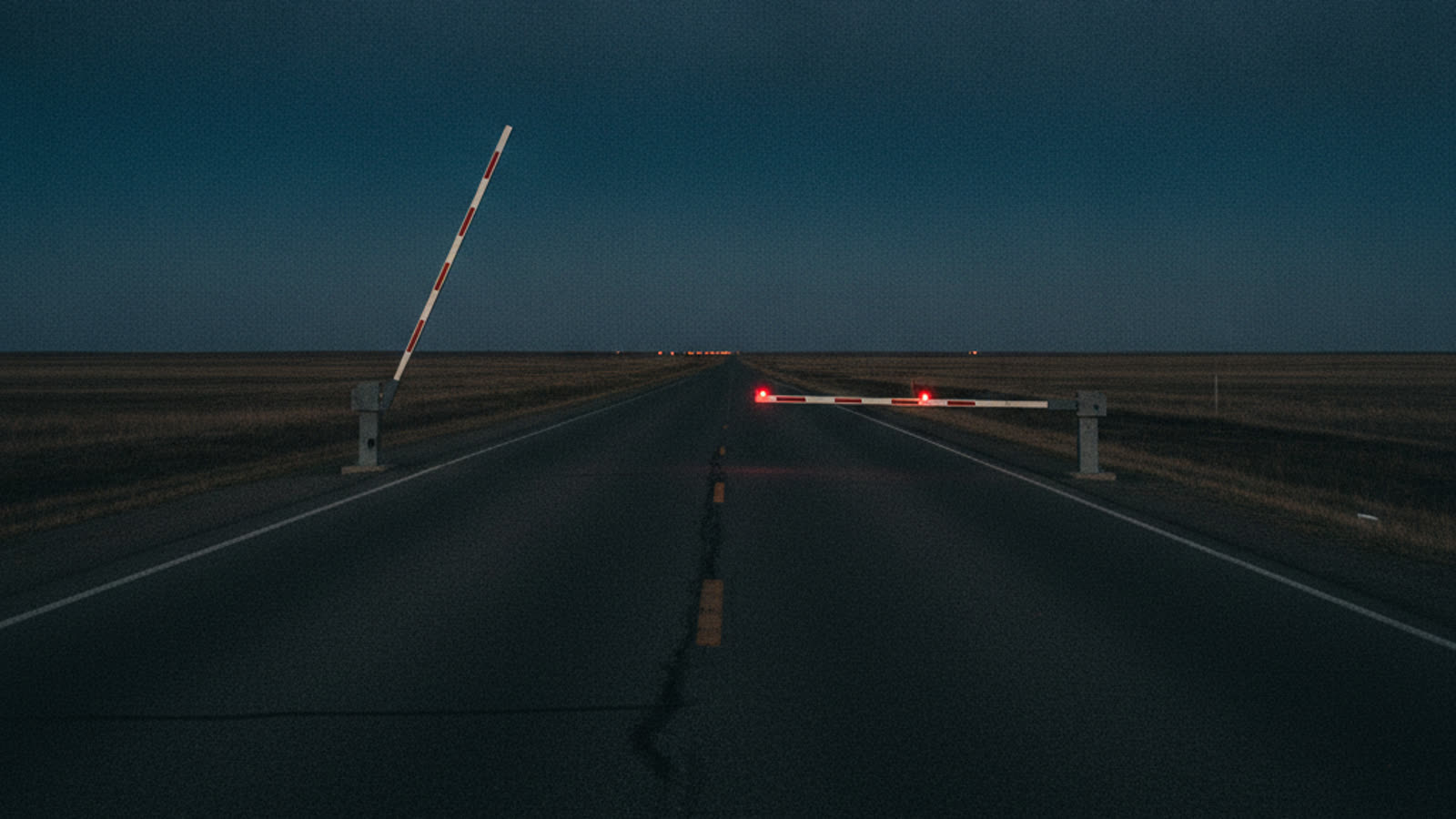 Una carretera recta y vacía al anochecer en una llanura abierta, con una barrera cercana levantada y otra lejana bajada e iluminada por una luz roja.