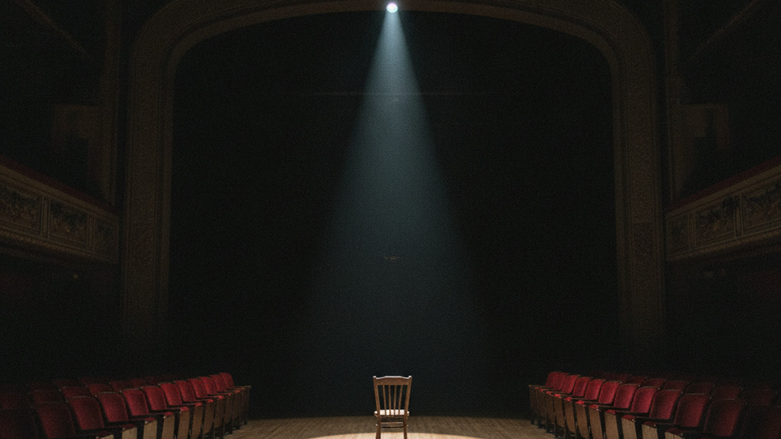 An empty theatre with red seats and one wooden chair on stage under a single spotlight.