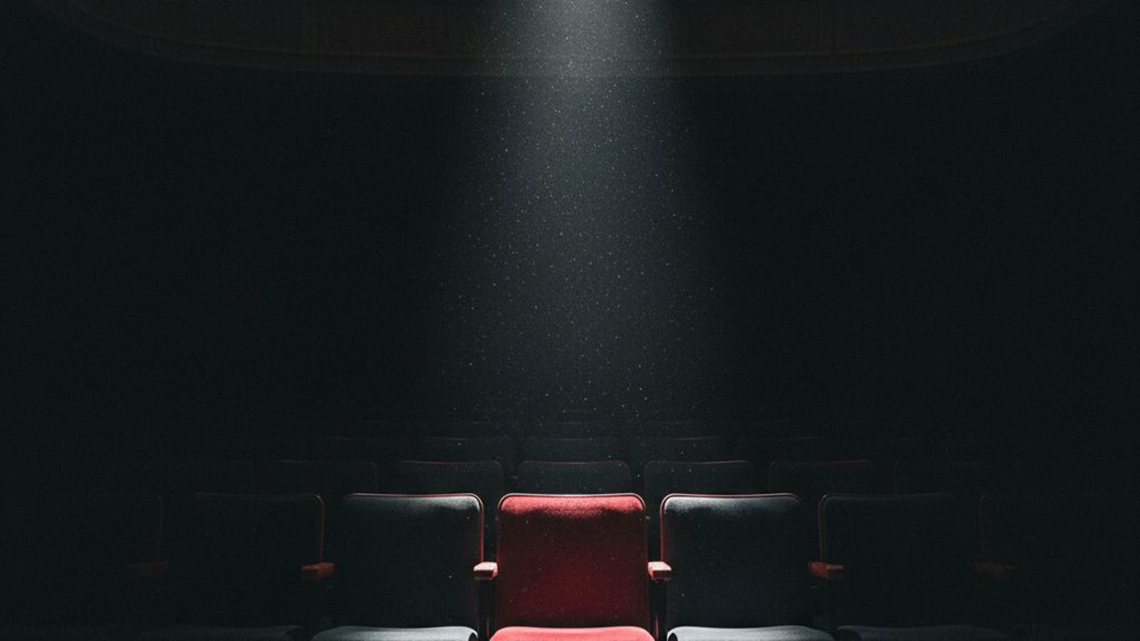 A row of empty theatre seats in the dark, a single spotlight falling on one empty red seat.